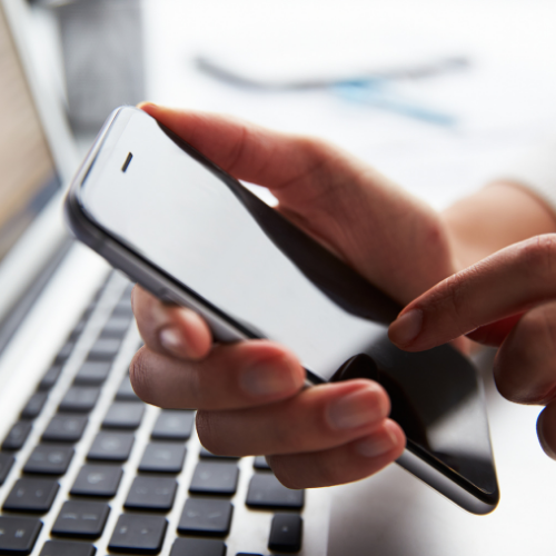 Hands typing on a phone with a laptop in the background